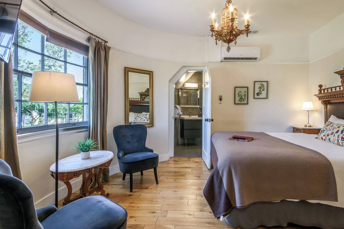 A hotel room with a bed, a chair, a small table with a plant, a chandelier, and a view of the bathroom.
