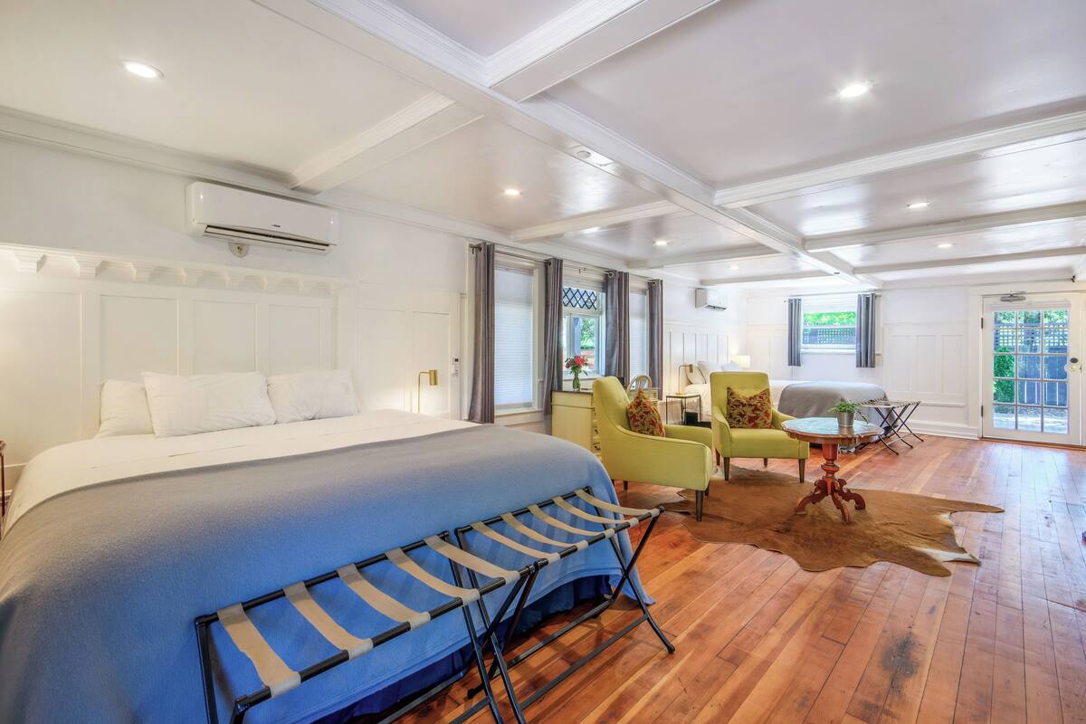 A spacious bedroom with a large bed, a seating area with a chair and a table, and a view of the outdoors through a door.