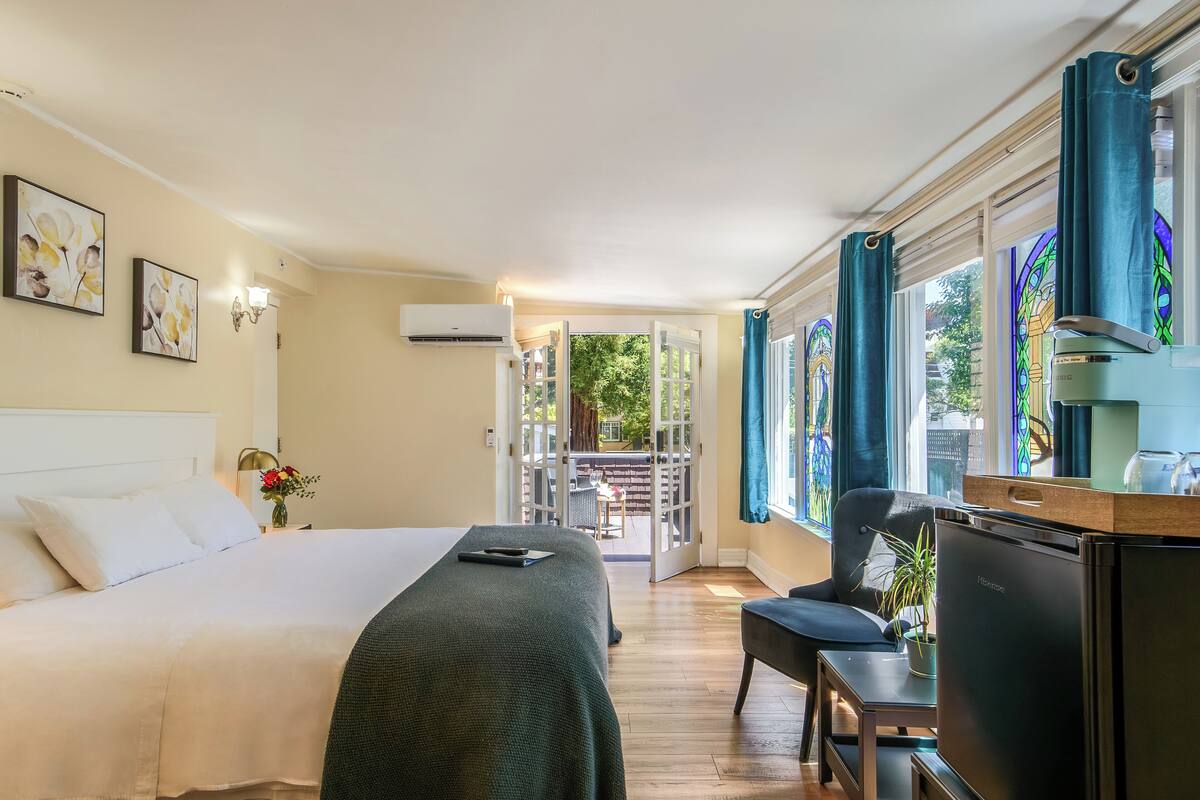 A hotel room with a bed, a desk, a refrigerator, and a view of an outdoor patio.