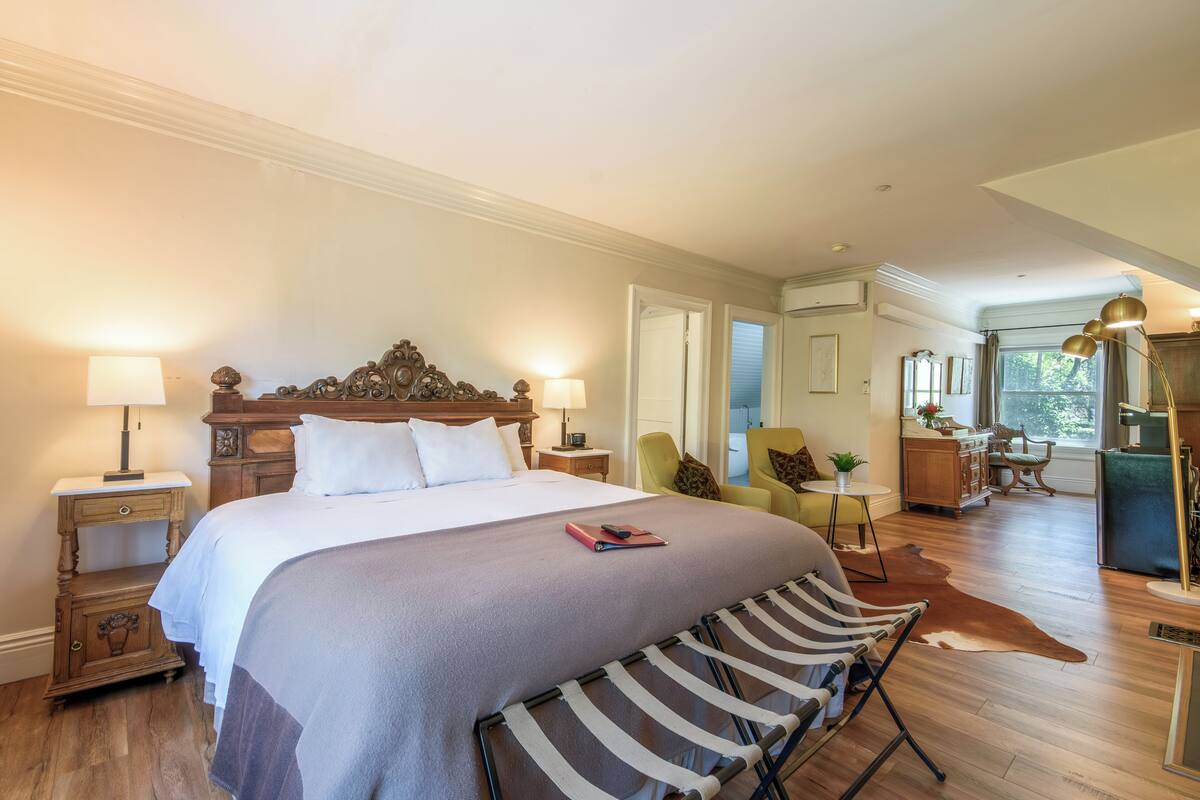 A bedroom with a large bed, bedside tables, a chair, a desk, and a view of the kitchen.