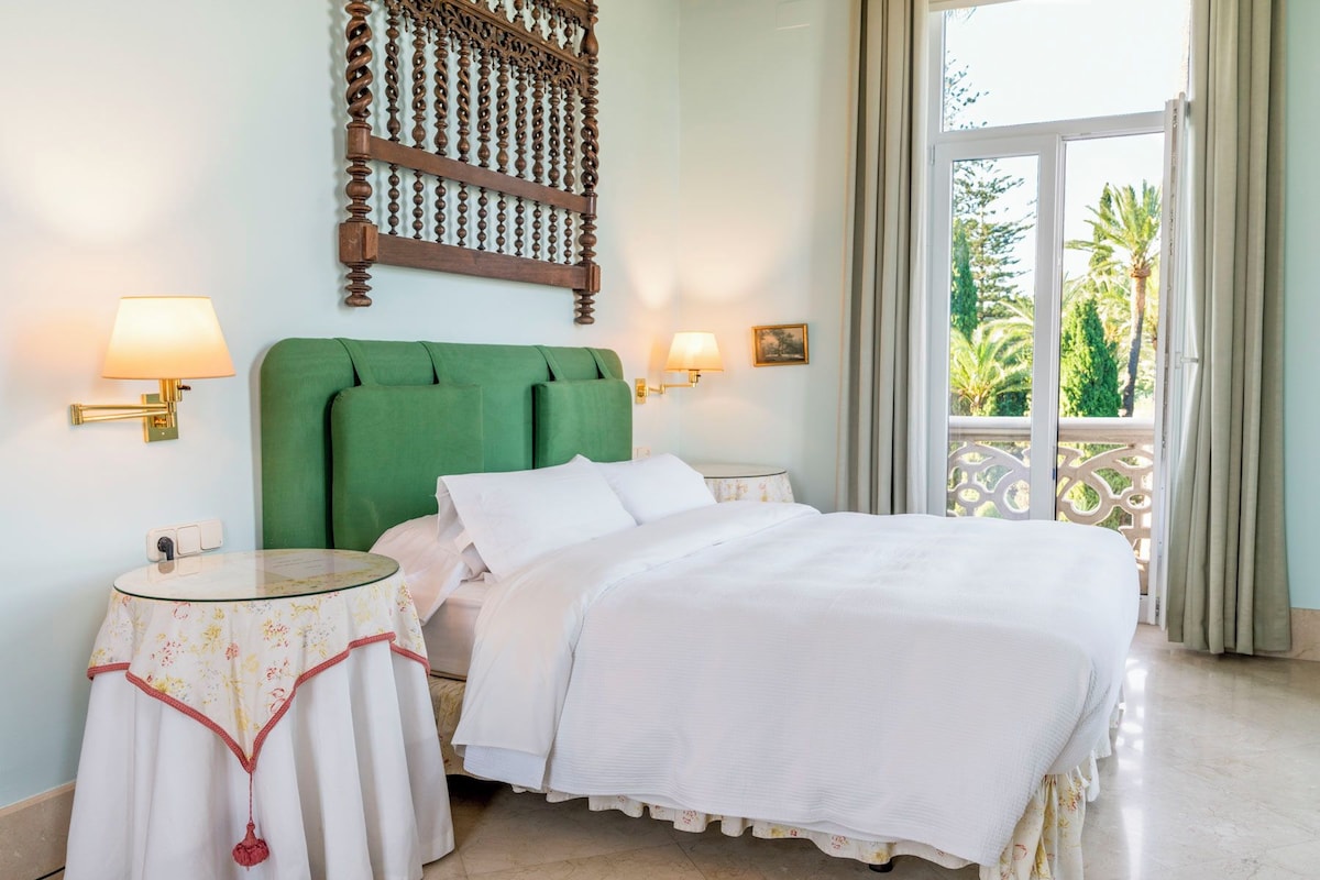 Habitación de hotel con cabecera tapizada en verde, dos mesitas de noche con lámparas, cama con ropa blanca y un balcón con cortinas.