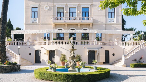 Un edifici gran i majestuós amb una entrada imponent, una font central i un jardí de disseny simètric.