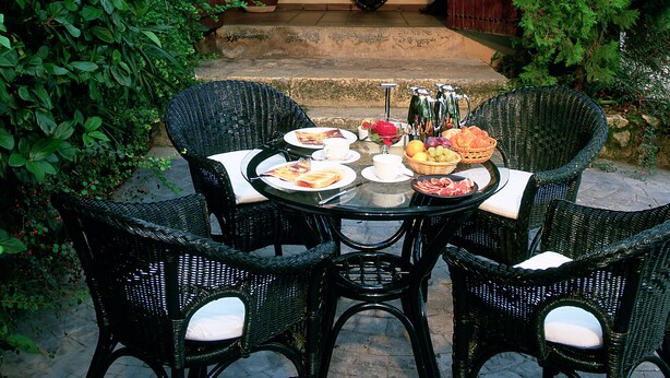 An outdoor dining area with wicker chairs and a table set for a meal, surrounded by lush greenery and a building with a tiled roof. - Rusticae Hotel LEstació (Bocairente)