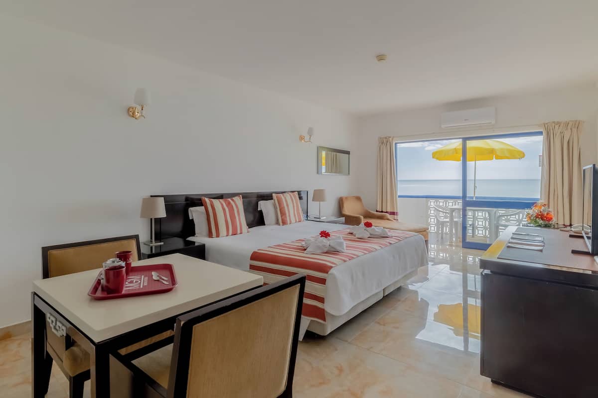A hotel room with a bed, a desk, a chair, a TV, and a balcony with a yellow umbrella.