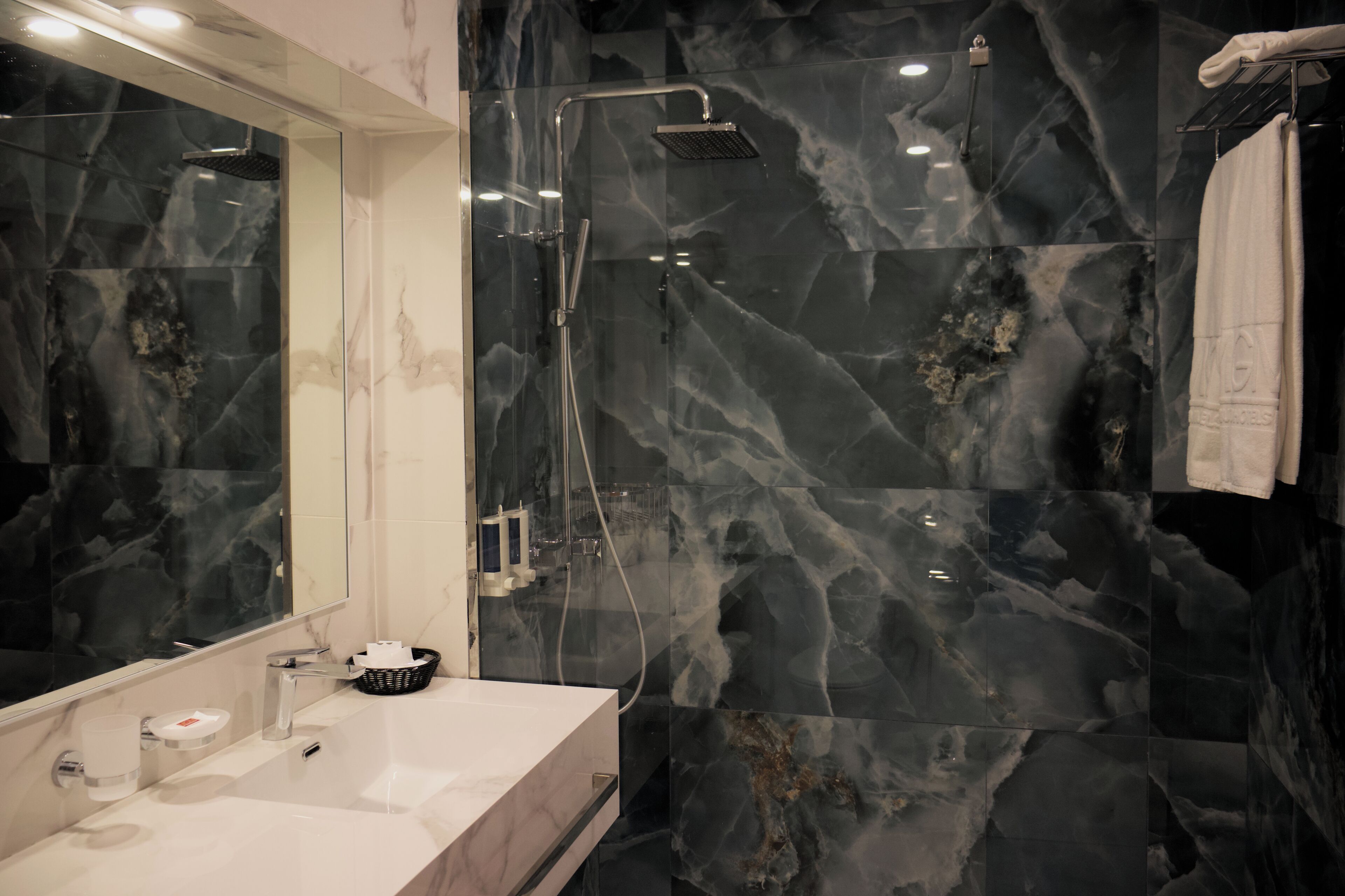 Une salle de bain moderne avec un grand miroir, un lavabo blanc, une douche équipée d’un pommeau de douche à main et des murs en marbre.