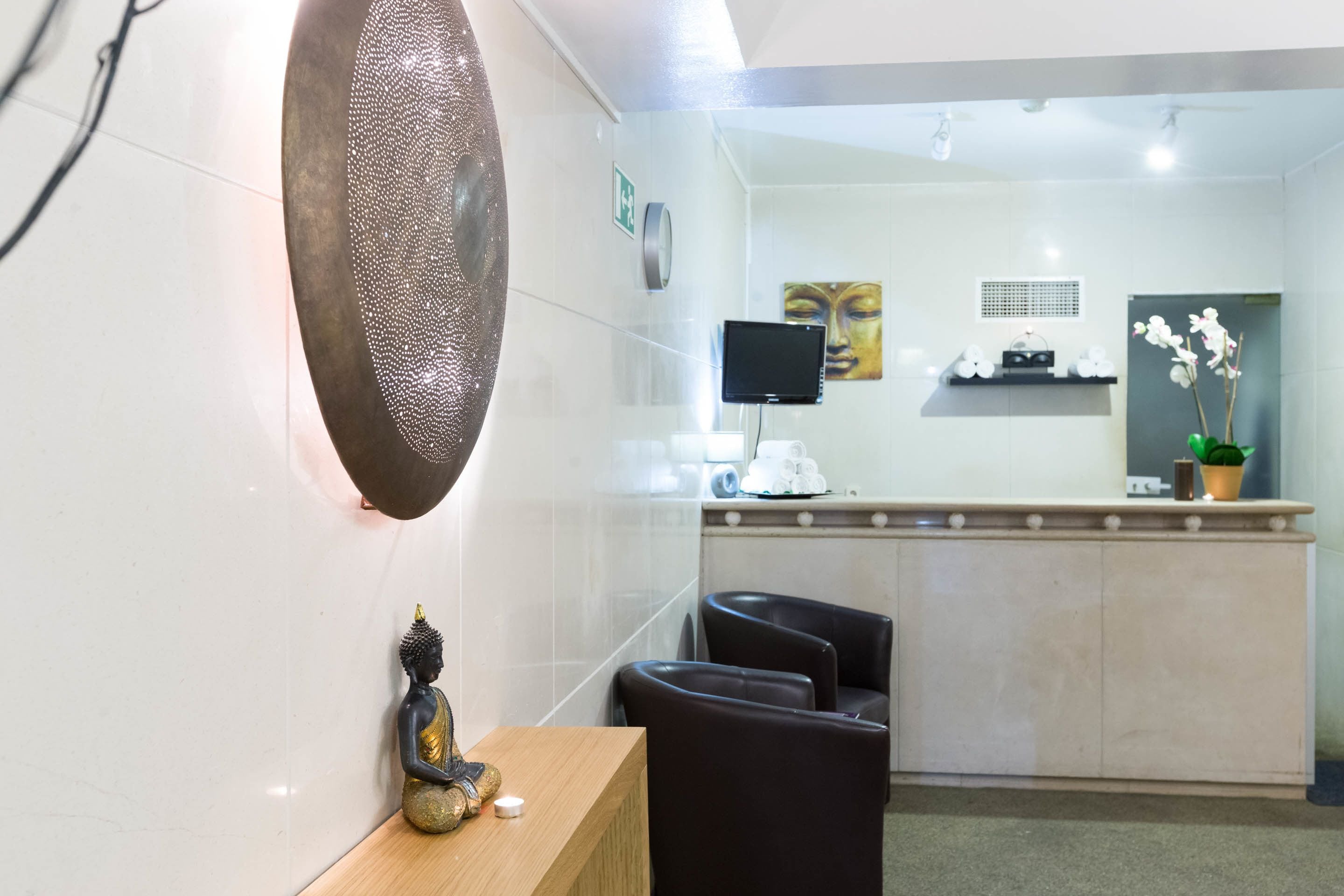 Un espace d’accueil comprenant un comptoir, des sièges, une statue de Bouddha, un écran fixé au mur et une plante en pot.