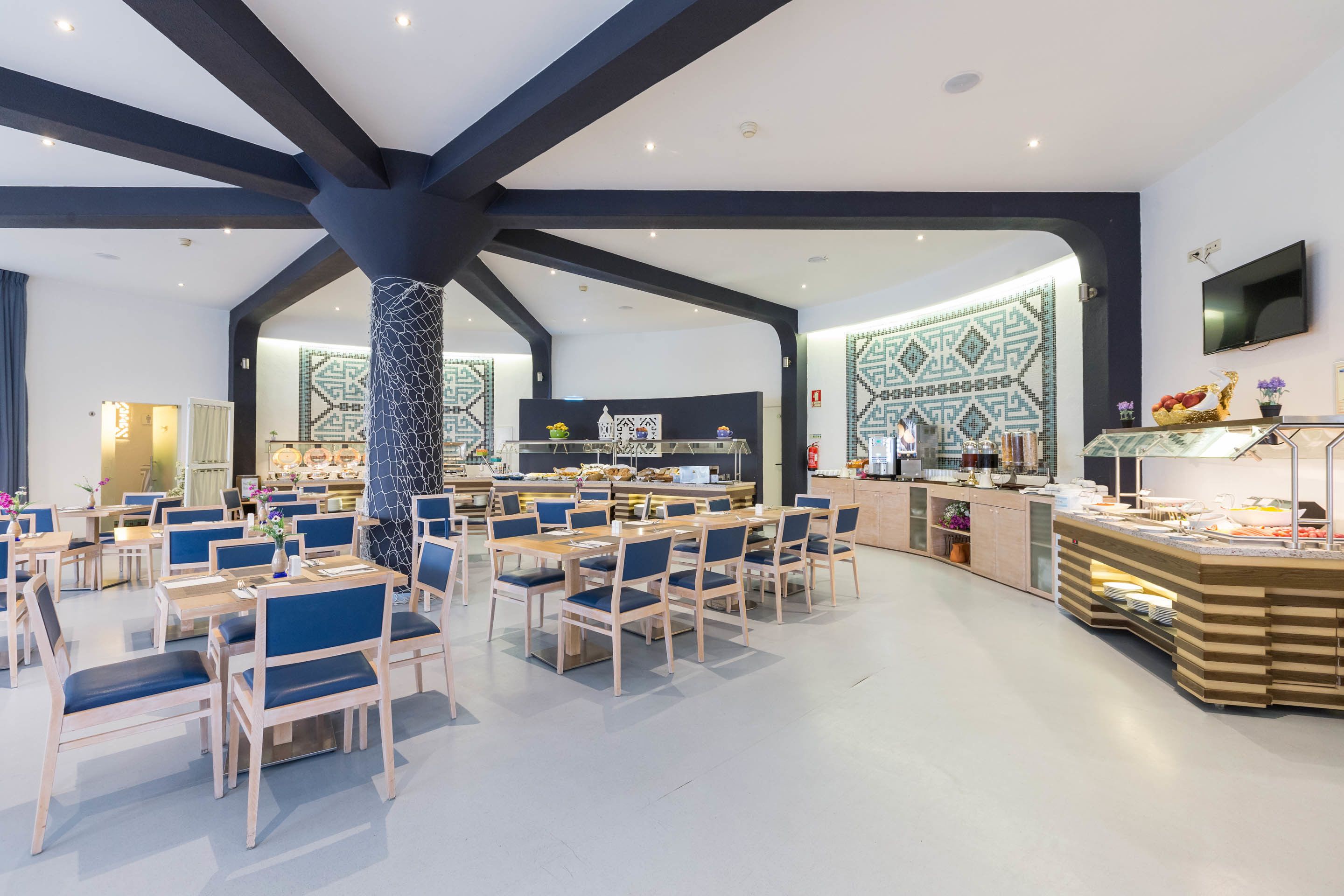 A spacious dining area with high ceilings and exposed beams, featuring a buffet setup with various food items and a television mounted on the wall.