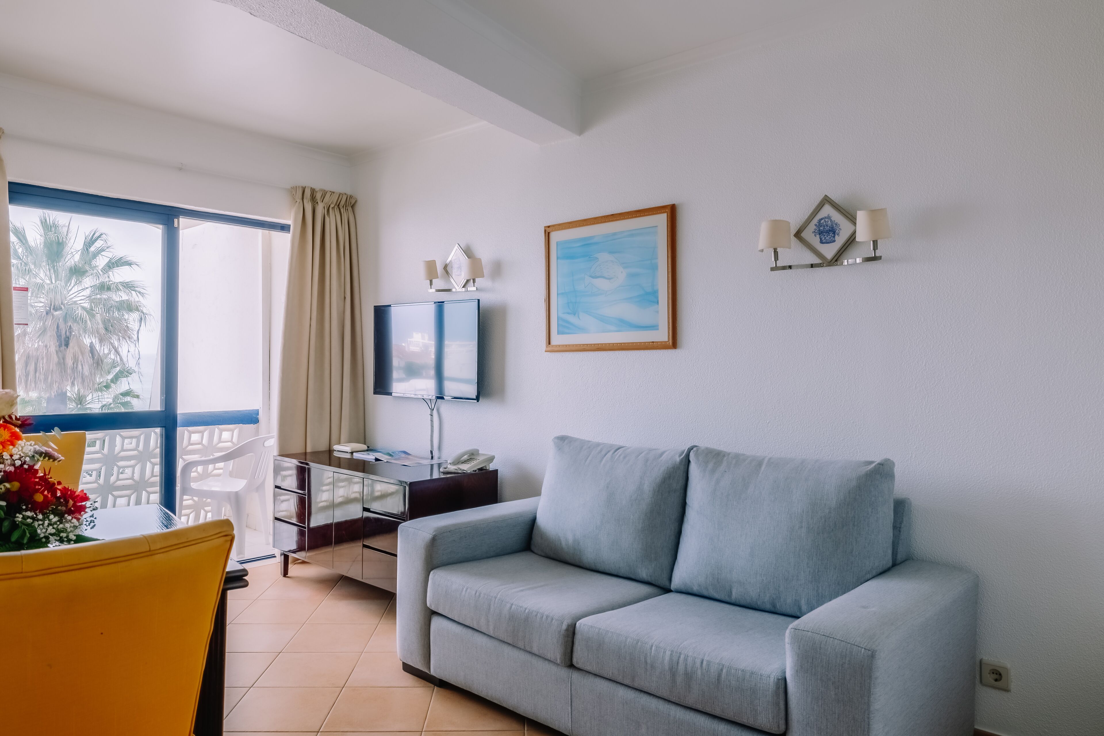 Un salon avec un canapé gris, un téléviseur posé sur un meuble, un tableau encadré au mur et un balcon avec vue sur un palmier.