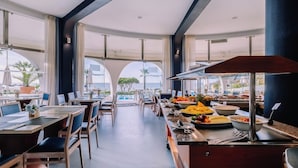 A modern dining area with tables and chairs, a buffet with fruits and vegetables, and large windows offering views of the outdoors. - Grand Muthu Oura View Beach Club (Albufeira)