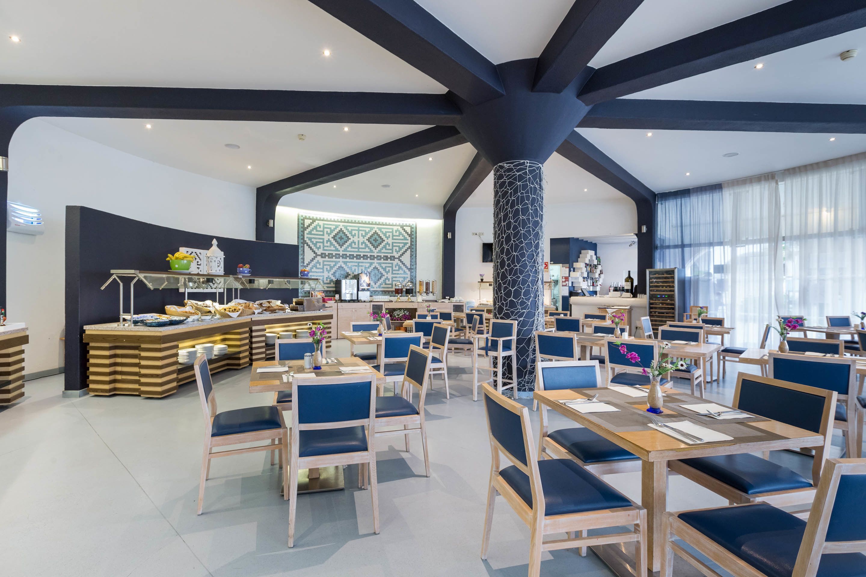A spacious dining area with multiple tables and chairs, a central pillar, and a buffet counter in the background.