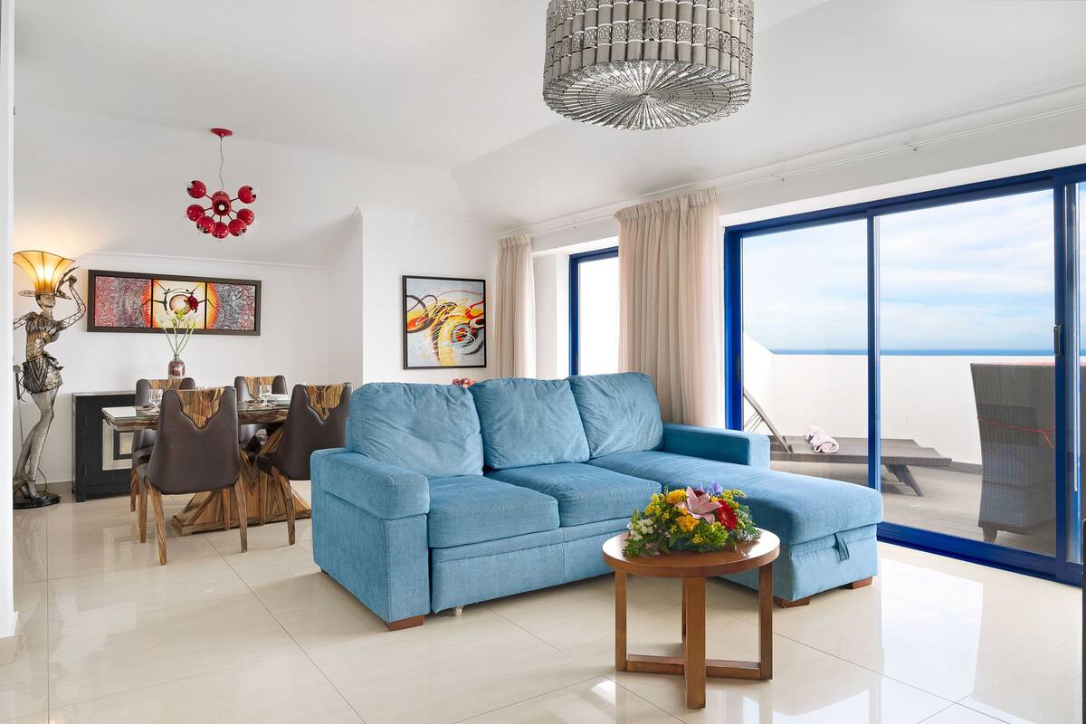 A modern living room with a blue sofa, dining table, and chairs, a decorative floor lamp, and artwork on the walls.