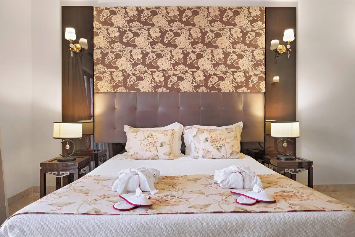 A hotel room with a large bed, floral-patterned headboard, two bedside tables with lamps, and towels on the bed.