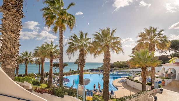 A pool area with palm trees, lounge chairs, and a view of the sea. - Grand Muthu Oura View Beach Club (Albufeira)