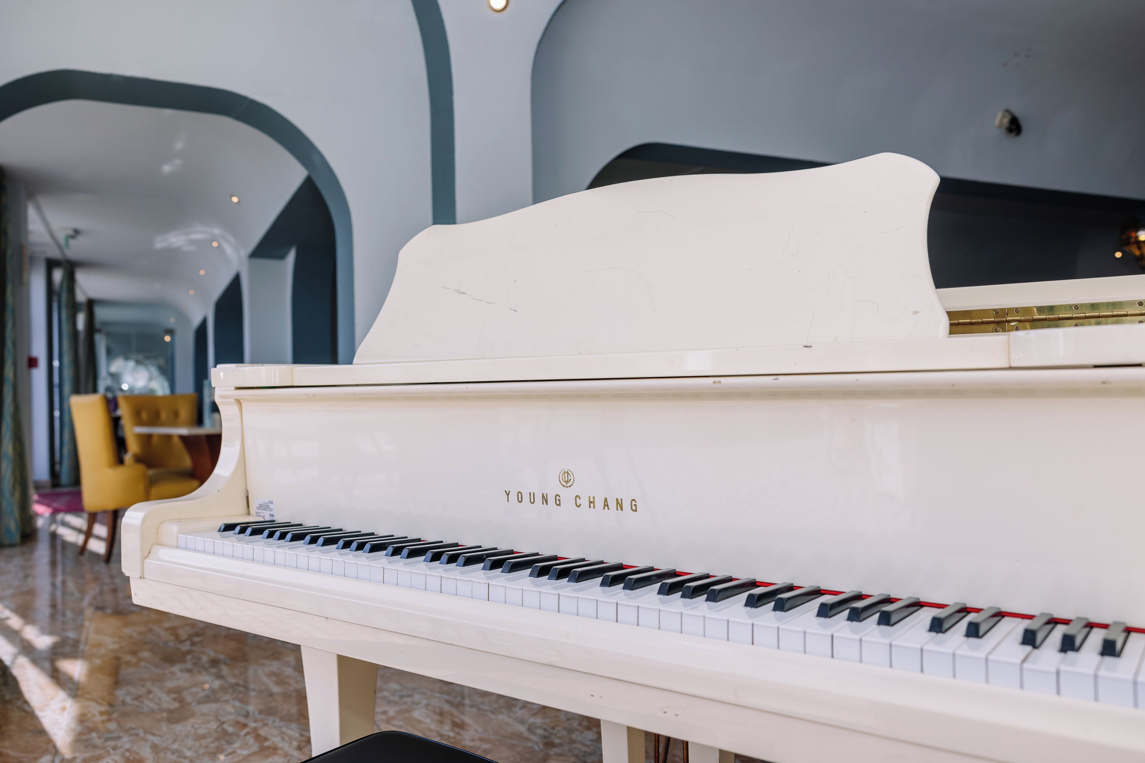 Un piano à queue blanc de la marque Young Chang, placé dans une pièce spacieuse et bien éclairée, dotée d’un sol en marbre et de chaises jaunes.