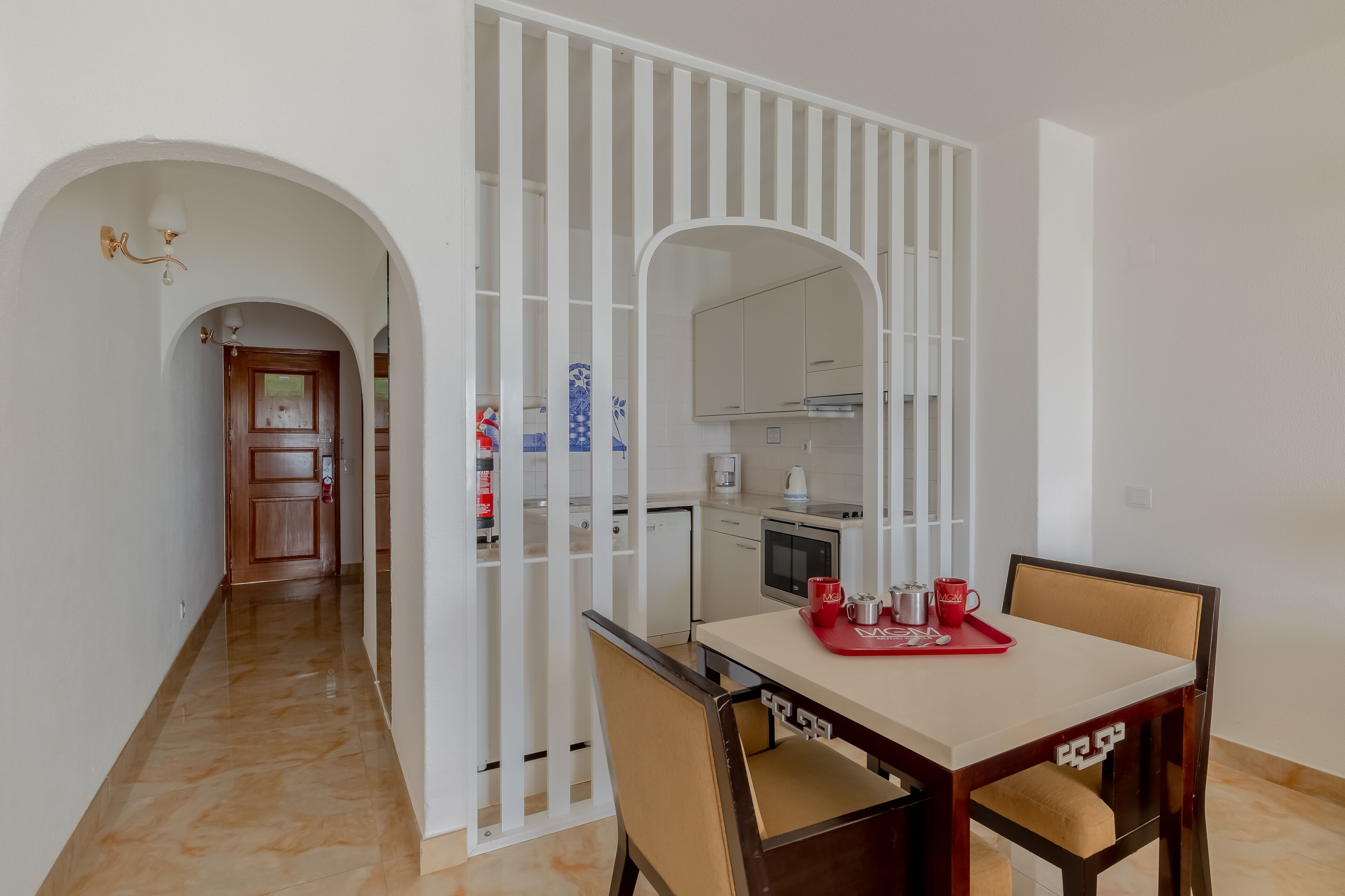 A dining area with a table and chairs, a kitchen in the background, and a white partition with vertical lines.