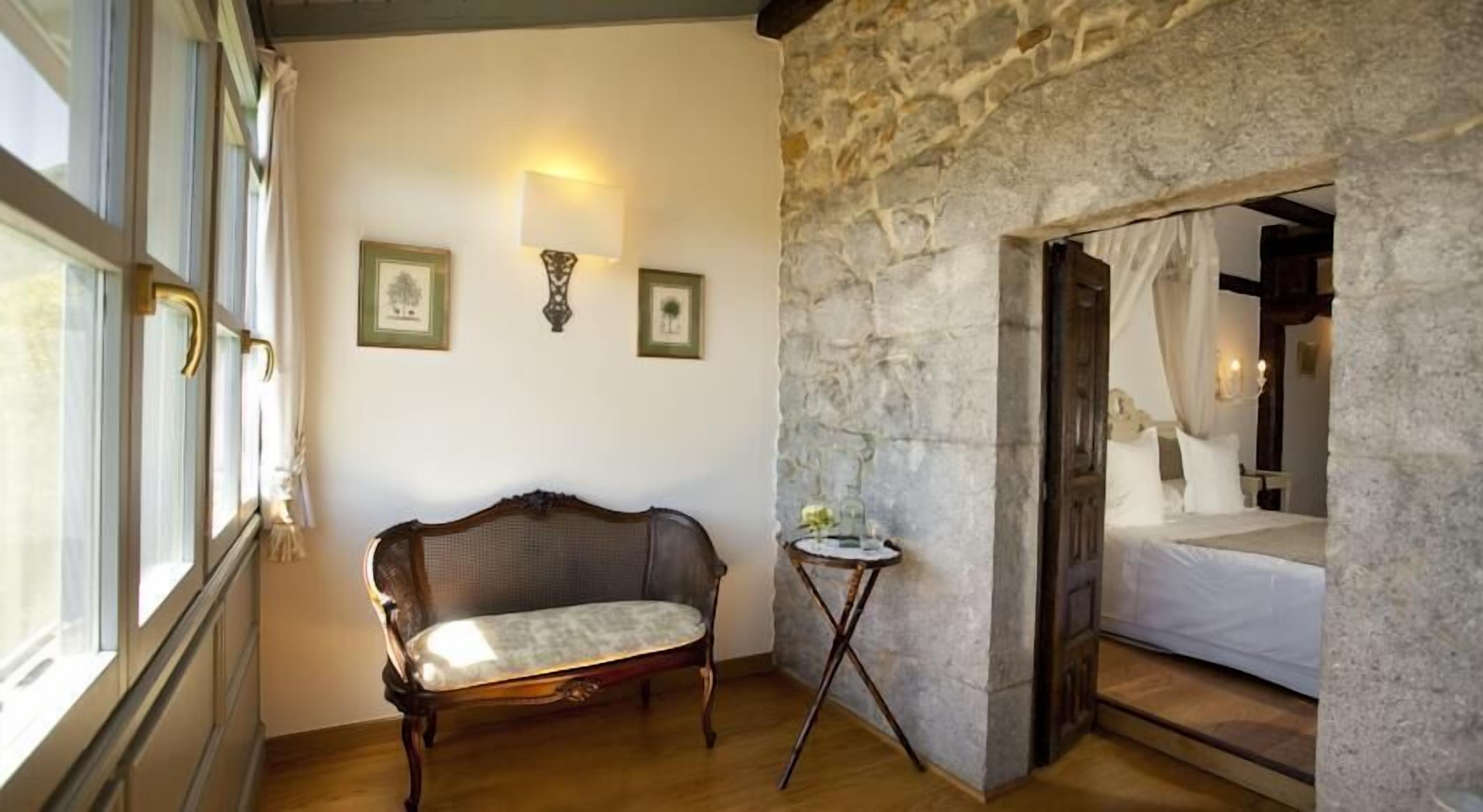 A room with a stone wall, a bed with white bedding, a wooden chair, and a small table with a lamp.
