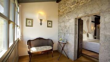 A room with a stone wall, a bed with white bedding, a wooden chair, and a small table with a lamp.