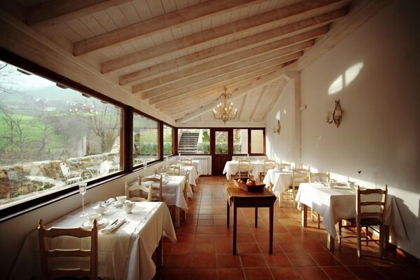 Un comedor con vigas de madera, mesas y sillas blancas, y ventanas amplias que dan a zonas verdes.