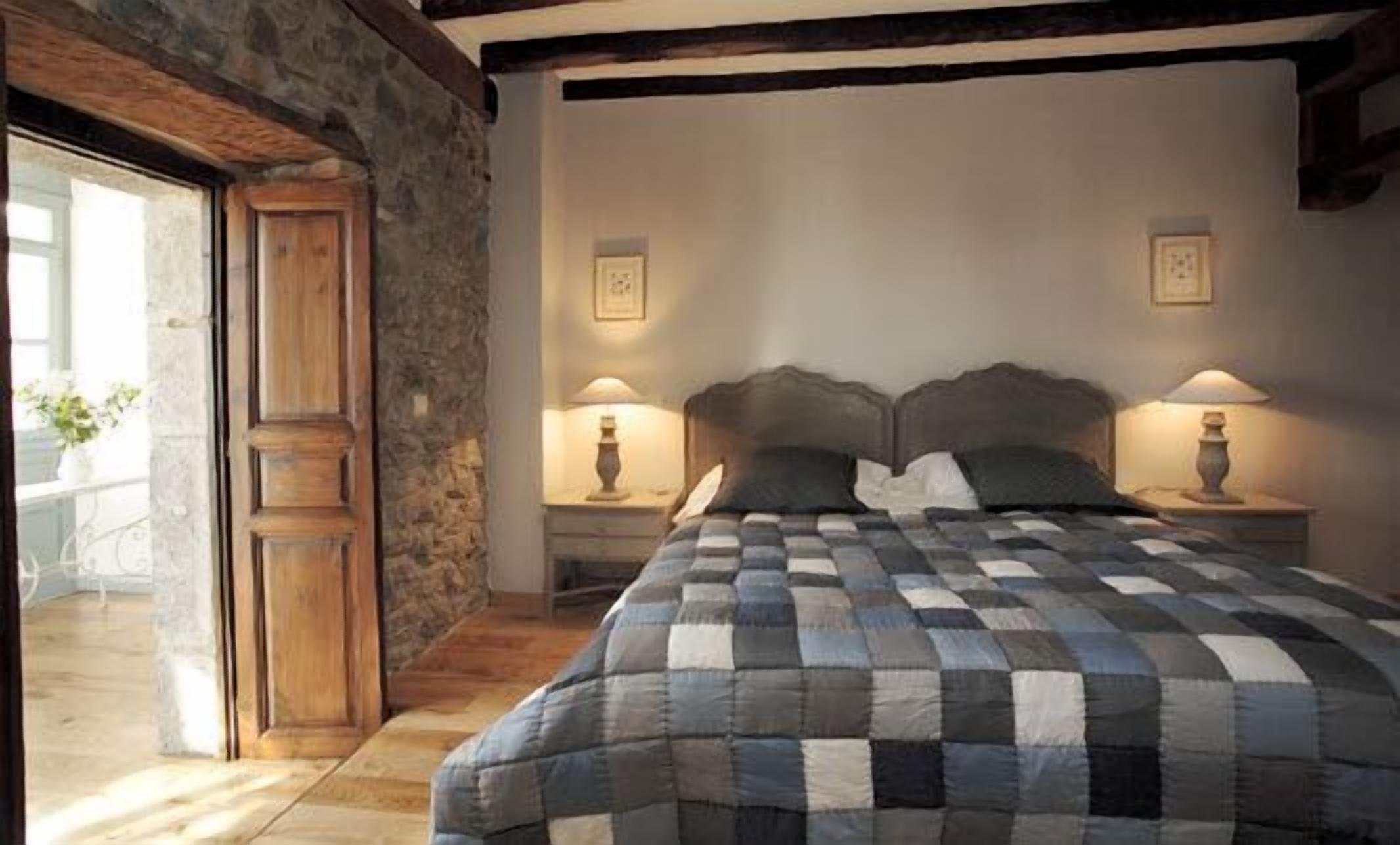 Une chambre à coucher avec une couvre-lit à carreaux, une tête de lit en bois, deux lampes de chevet et un mur en pierre.