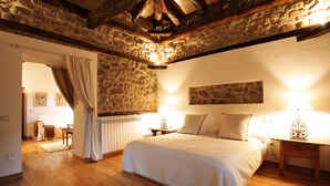 A bedroom with a stone wall, wooden beams, a bed with pillows, a nightstand, a lamp, and a radiator. - Hotel Casona de Quintana (Soba)