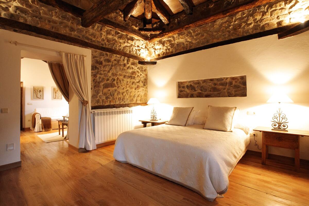 Ein Schlafzimmer mit Steinsandsteinwand, Holzbalken, einem Bett mit Kissen, einem Nachttisch, einer Lampe und einem Heizkörper.