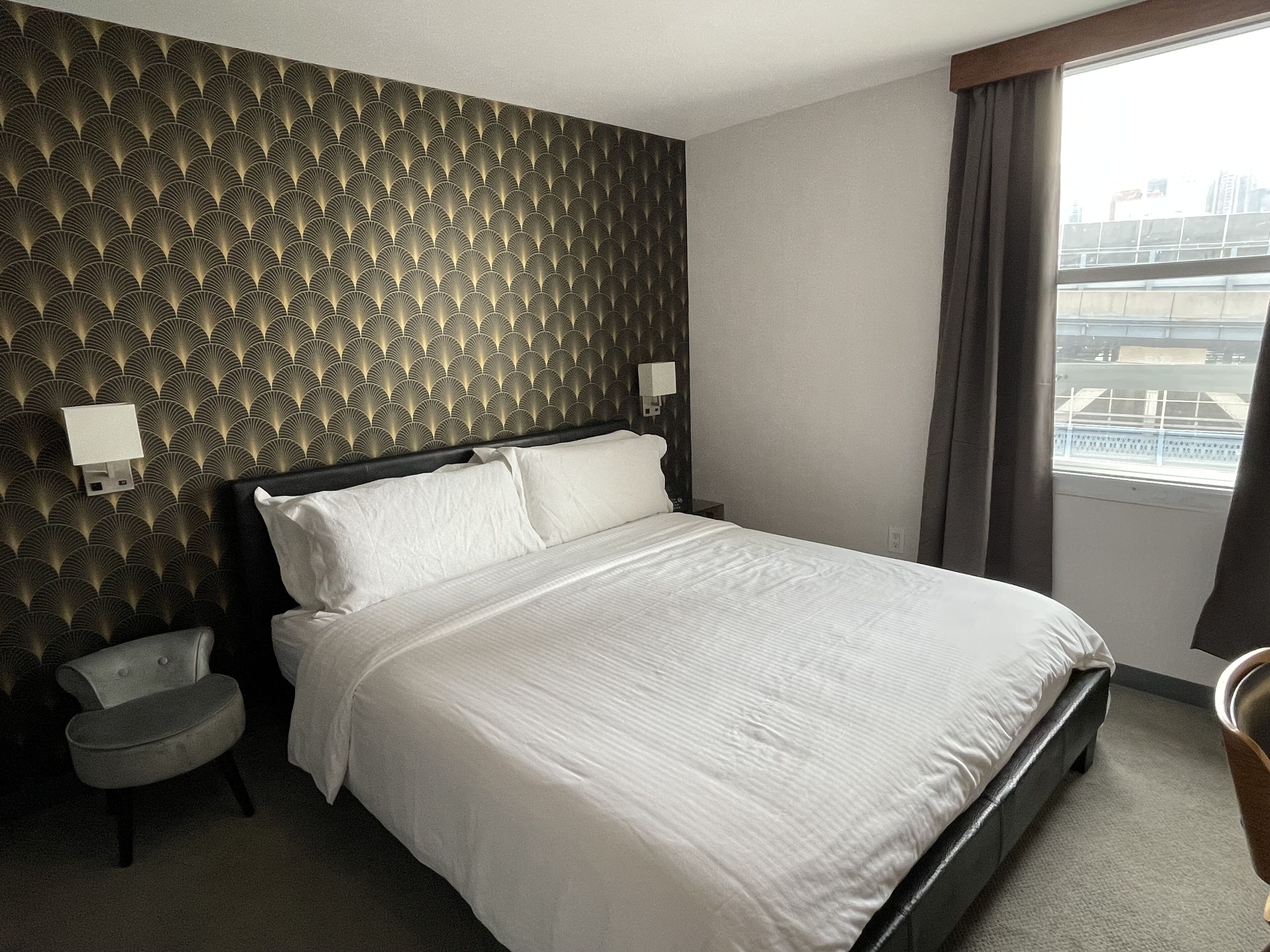 Habitación de hotel con cama, mesita de noche, silla, pared con diseño y ventana con vistas a edificios.