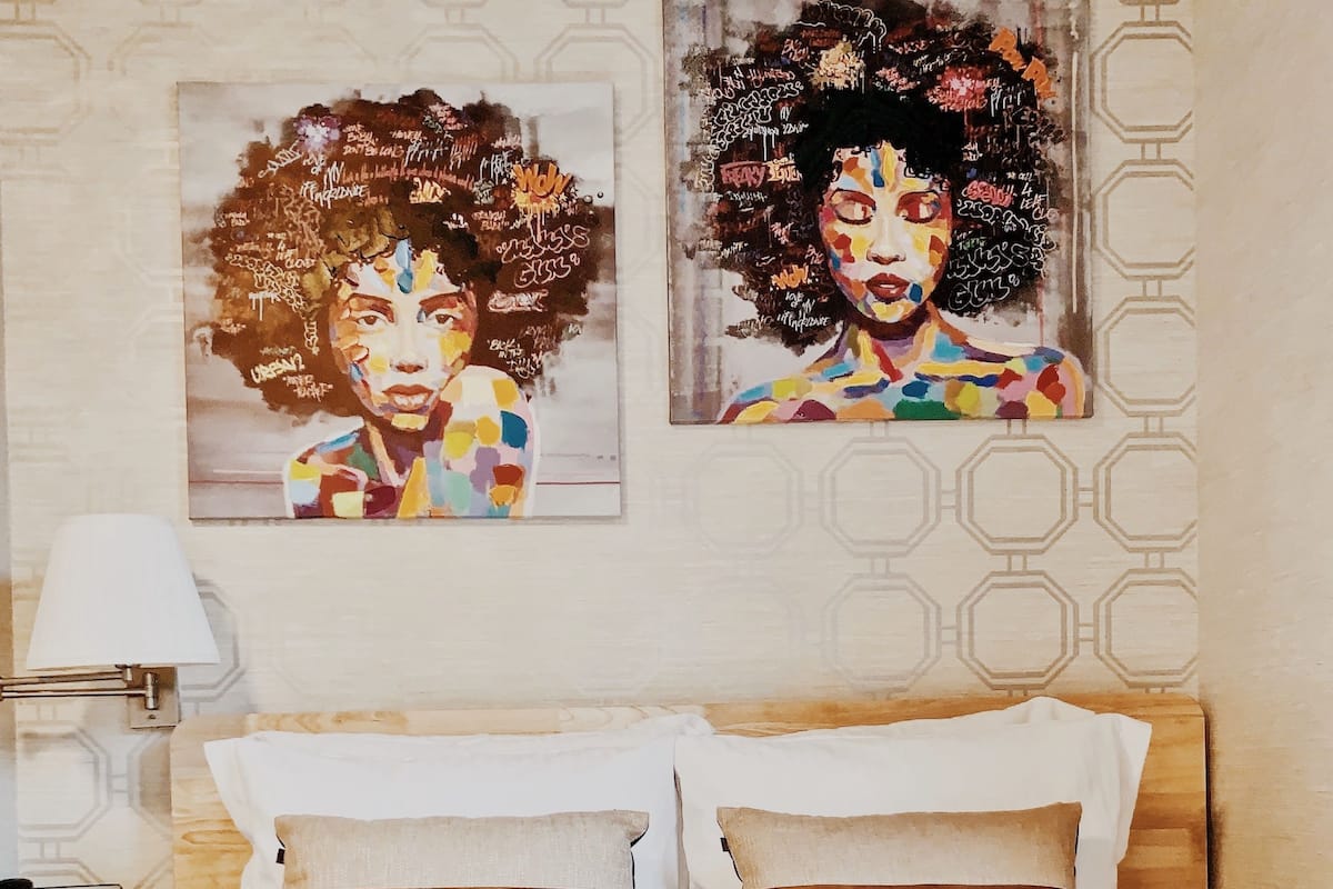 A hotel room with a bed, two pillows, a bedside table with a lamp, and two wall art pieces.