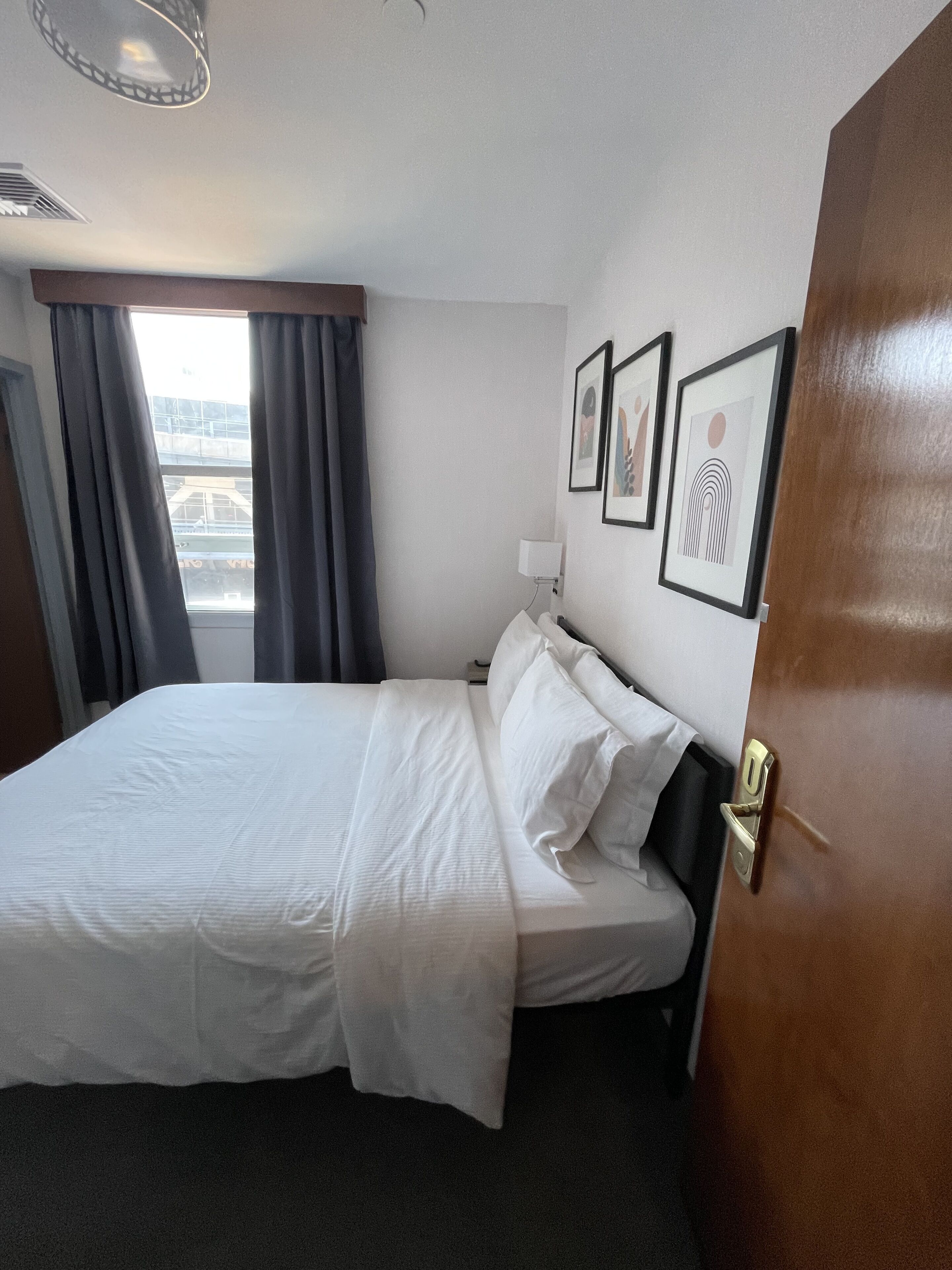 Una camera d'albergo con un letto rifatto, un comodino con una lampada e una finestra con tende. Alle pareti ci sono quadri incorniciati.