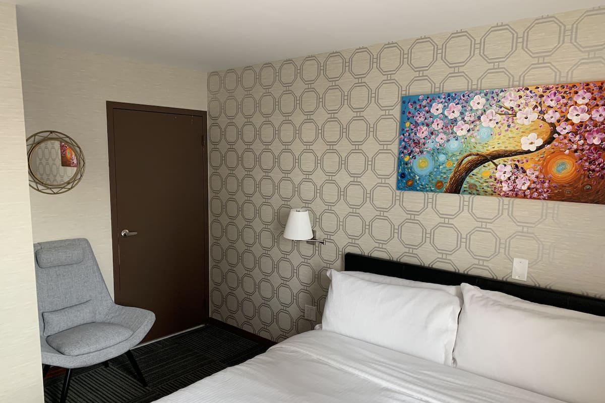 A hotel room with a bed, a chair, a door, a wall with a geometric pattern, and a colorful painting.