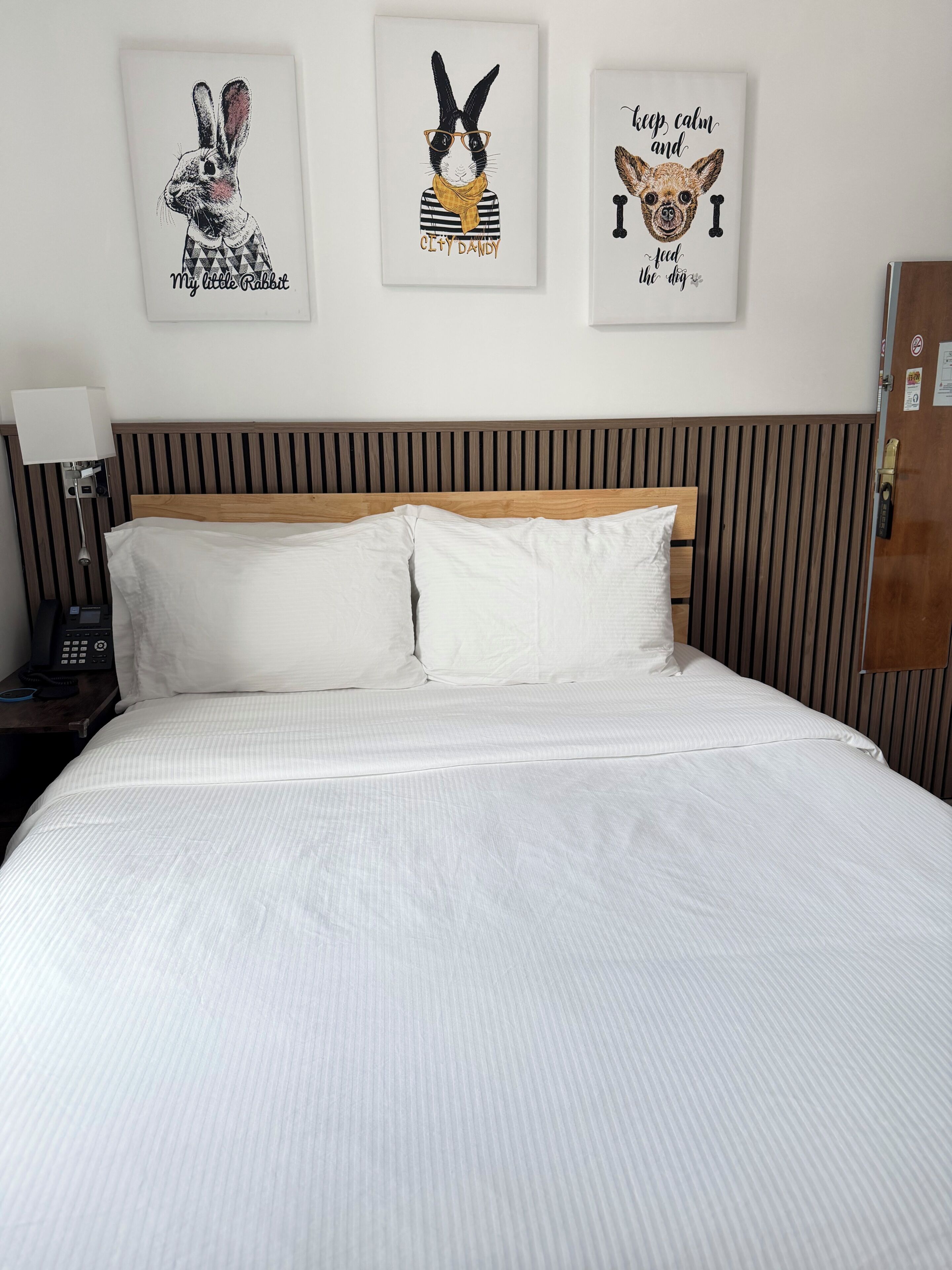 A neatly made bed with white linens, a wooden headboard, and three framed artworks above it.
