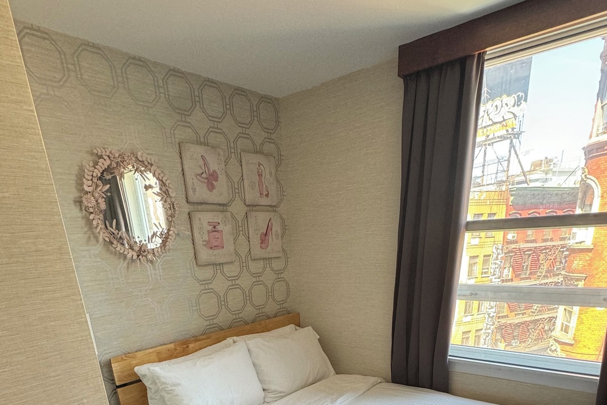 A hotel room with a bed, a mirror, and a window with a view of buildings.