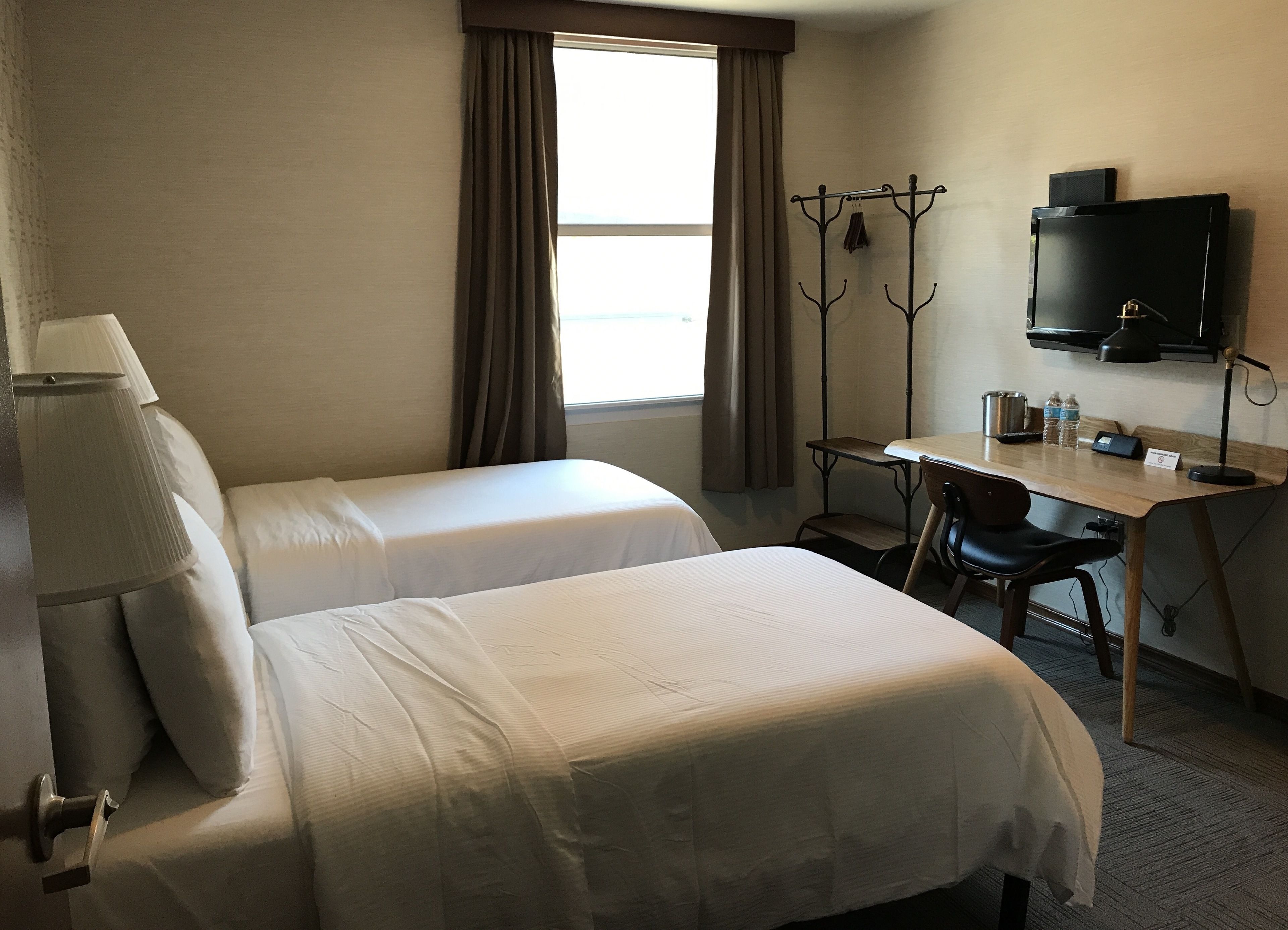 A hotel room with two beds, a desk, a chair, a TV, and a coat rack.