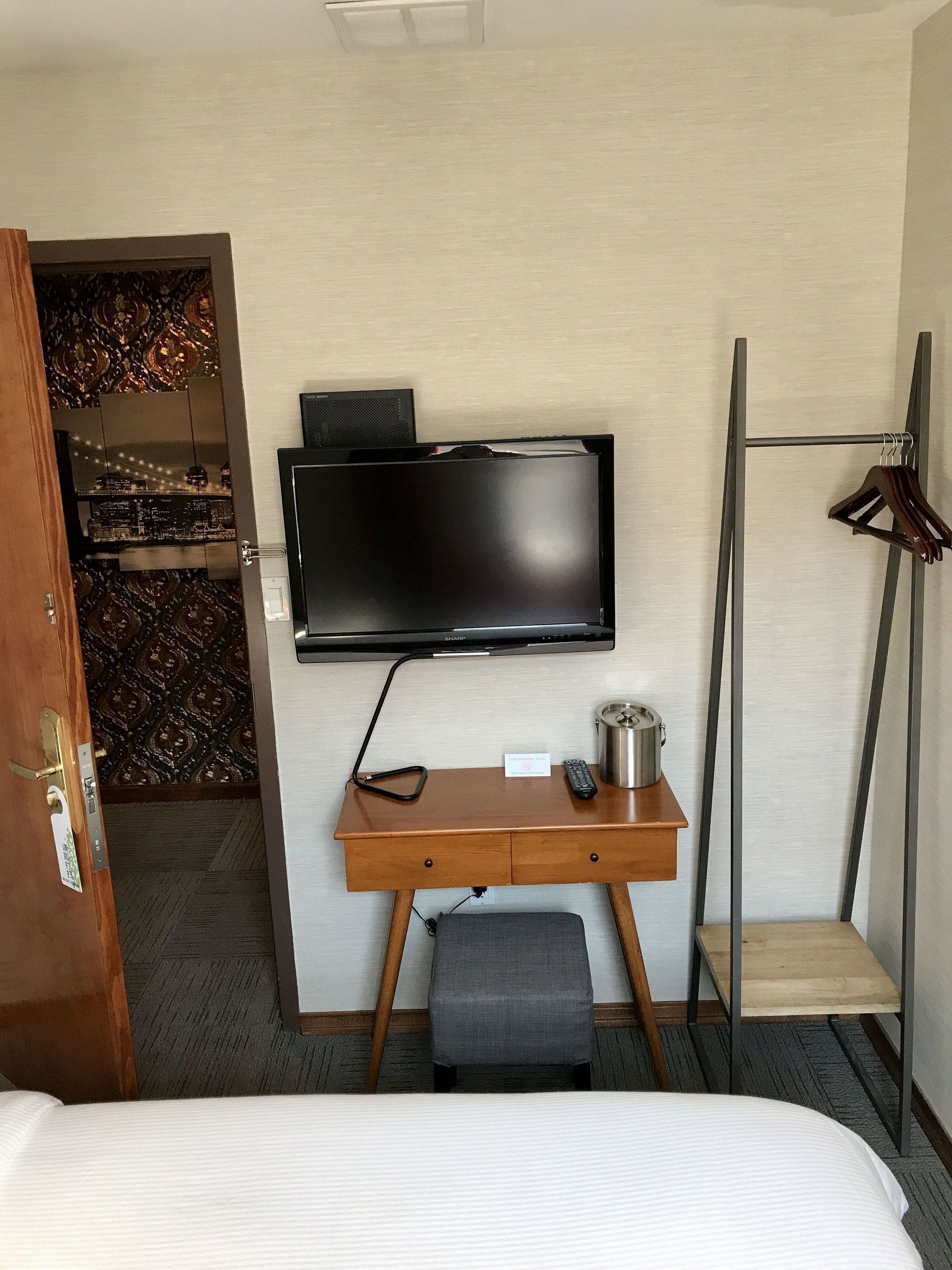 A hotel room with a bed, a desk with a flat-screen TV, a small stool, a coat rack, and a door leading to a patterned room.