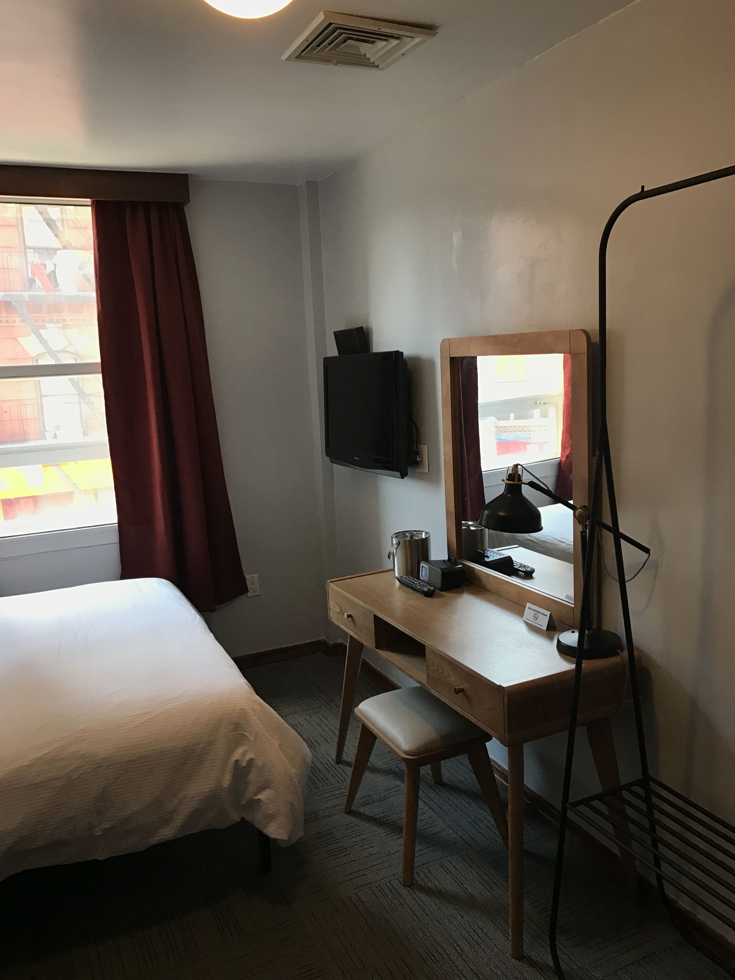A hotel room with a bed, a desk with a chair, a television mounted on the wall, and a standing lamp.