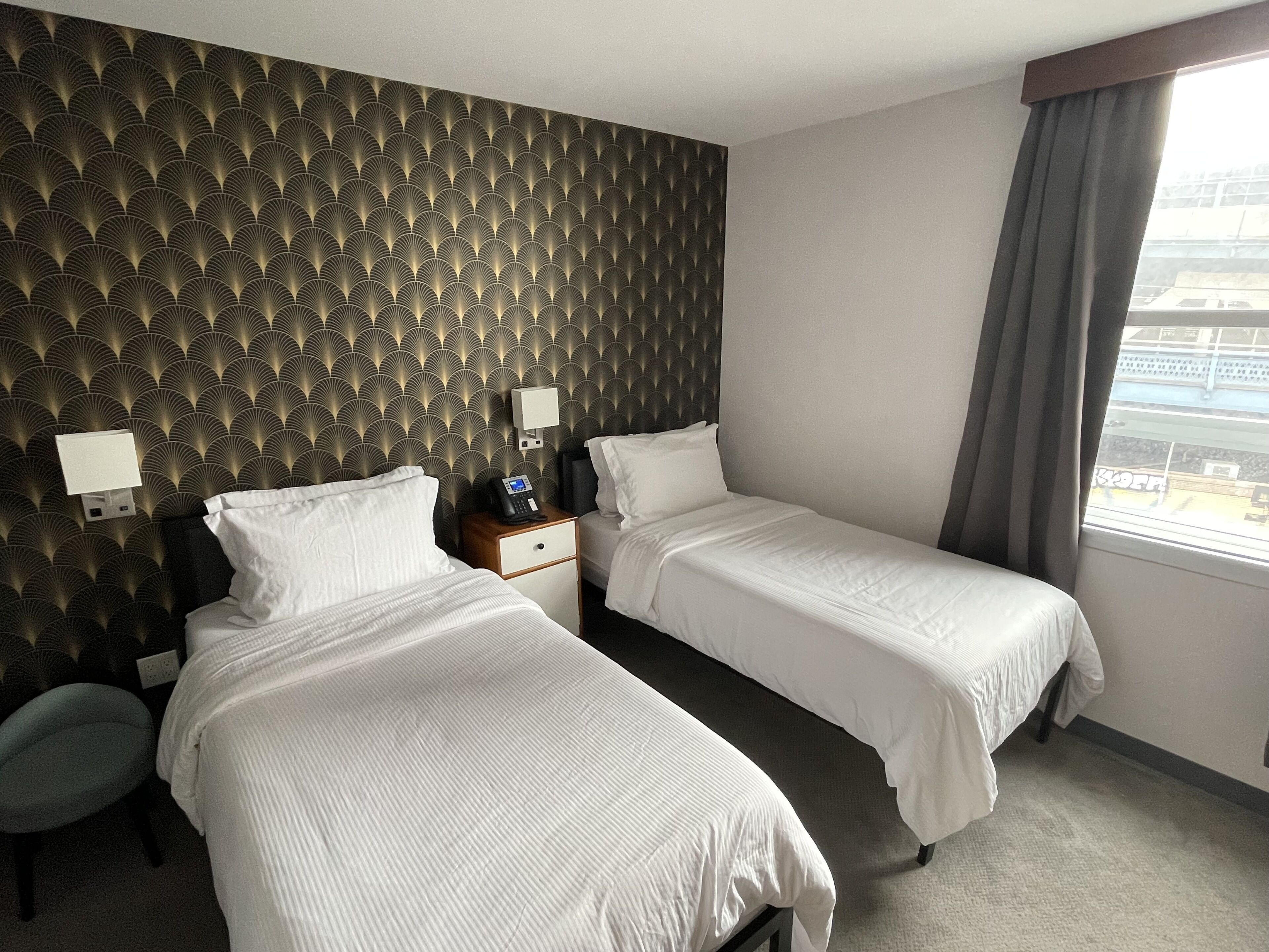 Habitación de hotel con dos camas, una mesita de noche con teléfono, una lámpara de pared y una ventana con cortinas.