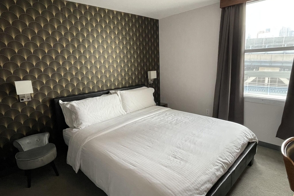 A hotel room with a bed, a chair, a nightstand, a wall with a patterned design, and a window with curtains.