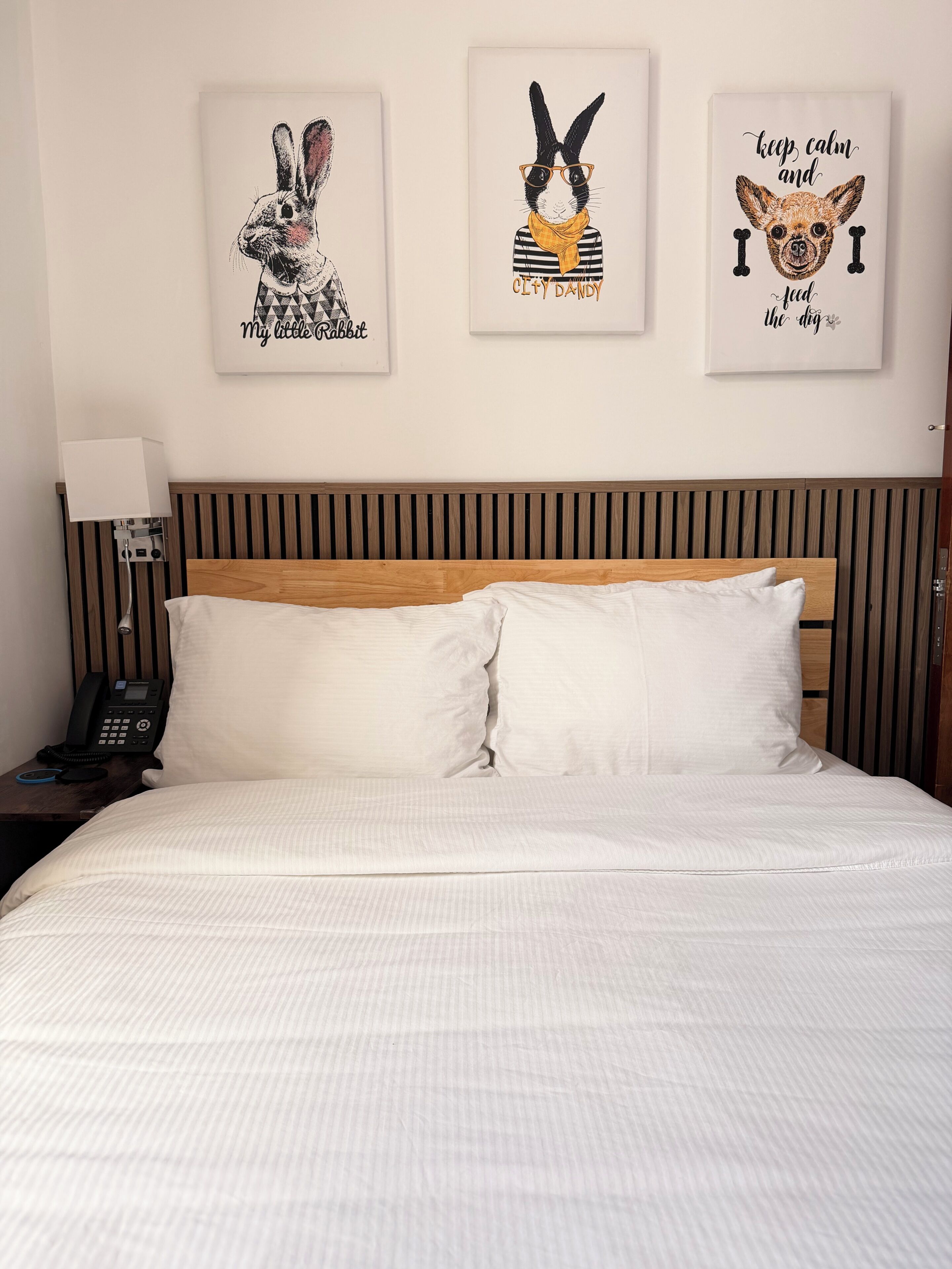 Una cama bien tendida con sábanas blancas, cabecera de madera y tres cuadros enmarcados en la pared superior.