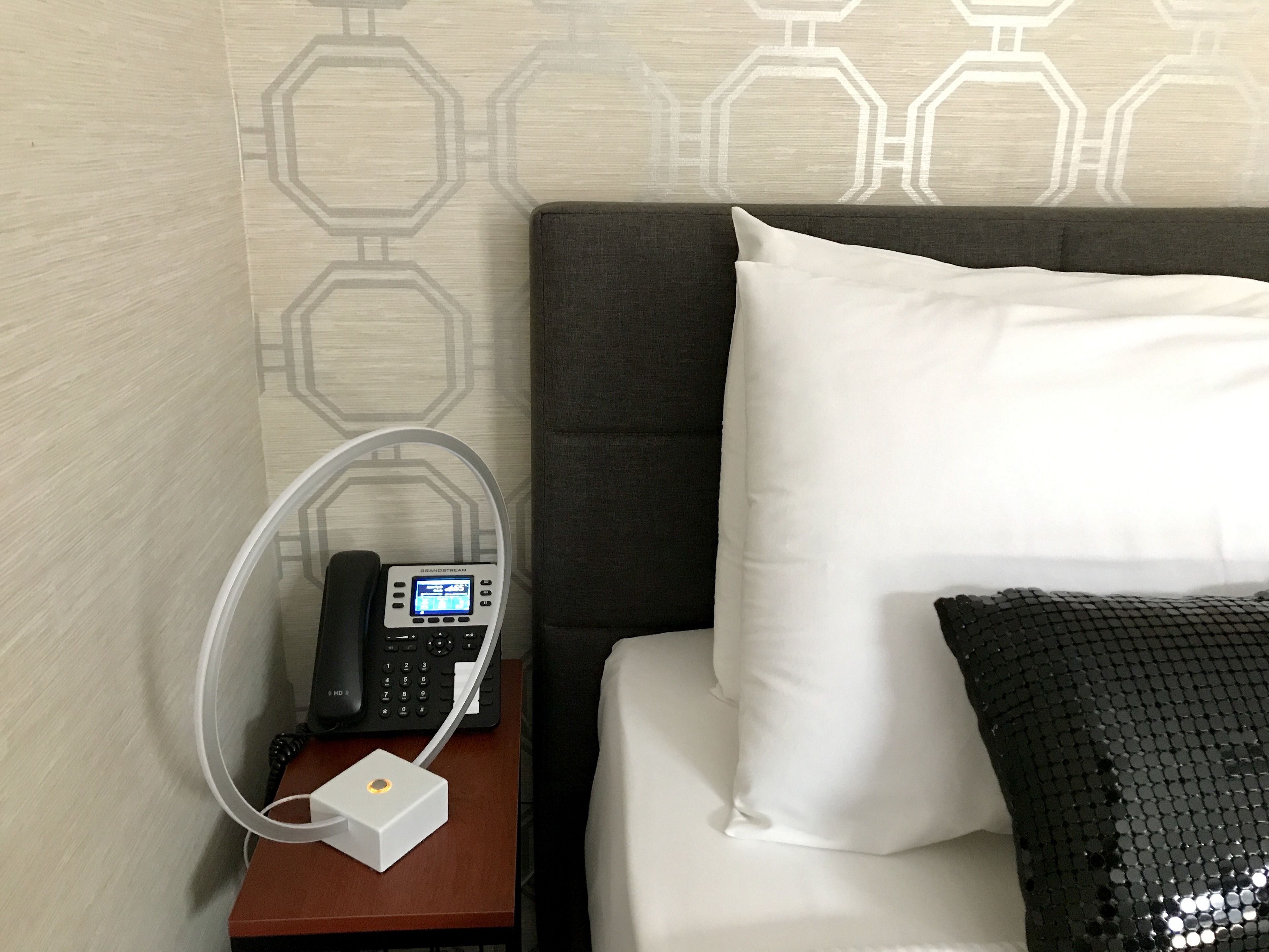 A hotel room with a bed, a bedside table with a phone and a charging station, and a patterned wall.