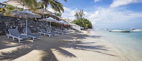 Een strand met witte ligbedden en parasols, een stenen muur en boten in het water.
