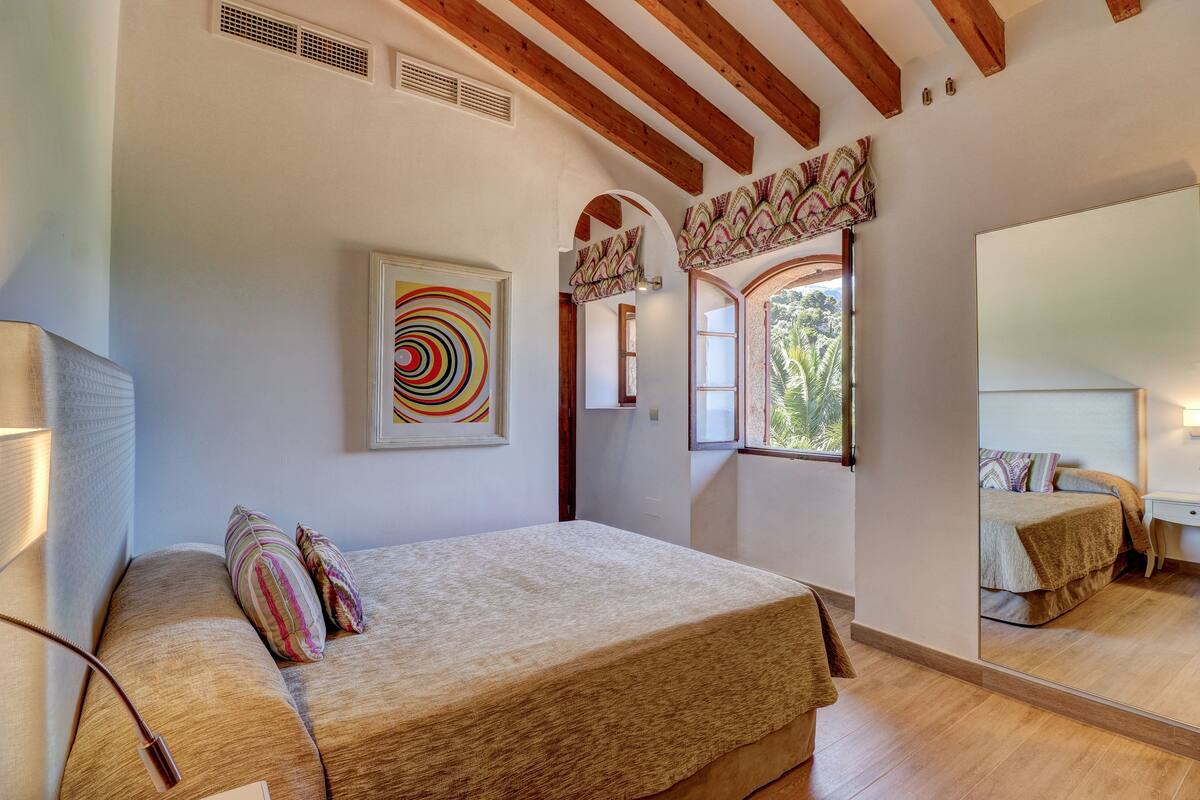 Une chambre avec un grand lit, un miroir, une fenêtre donnant sur un beau paysage et un plafond avec des poutres apparentes.