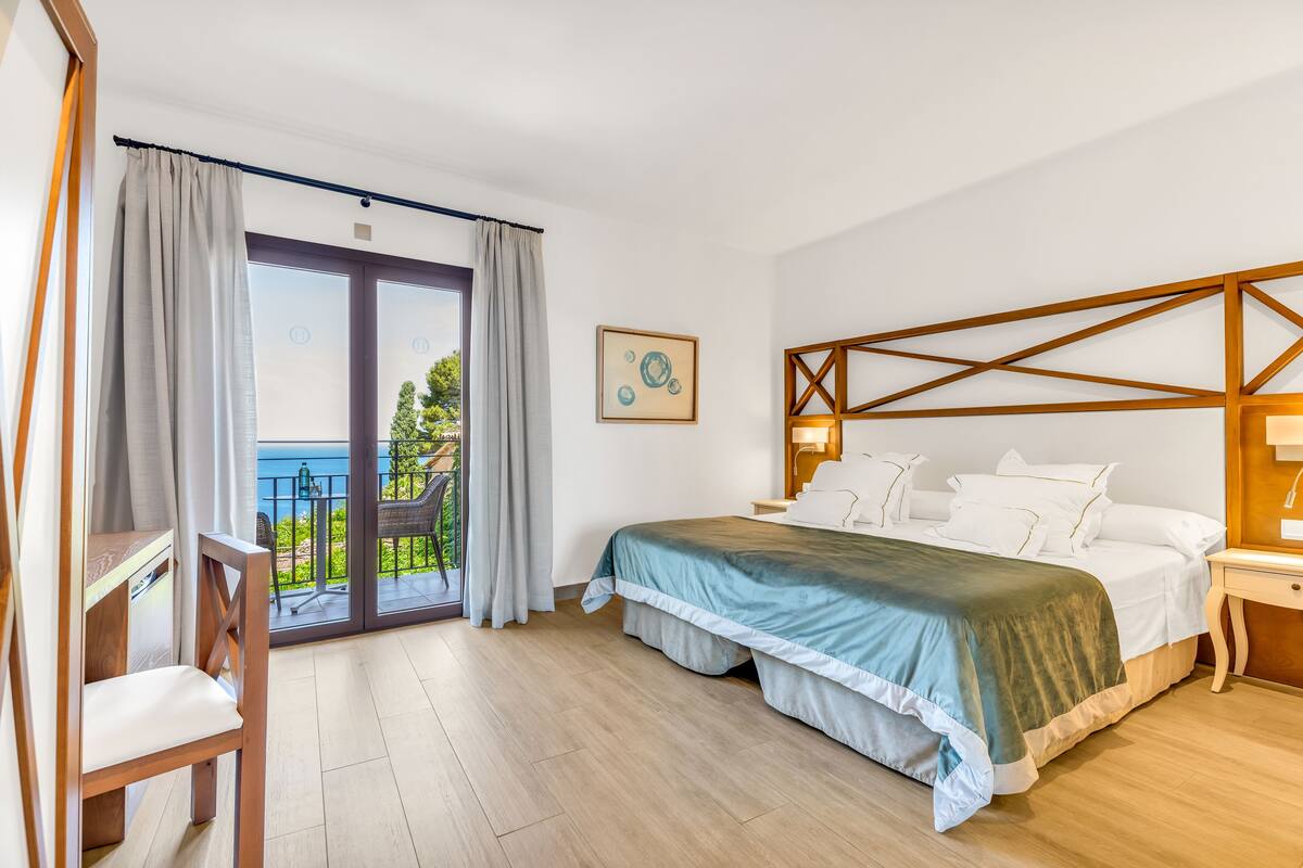 Une chambre à coucher comprenant un lit, une chaise, une table de chevet, une lampe, un tableau au mur et une vue sur l’extérieur.