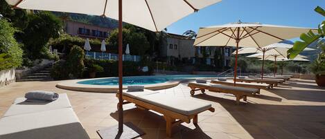 Zona junto a la piscina con sillones de descanso y sombrillas, una piscina y un edificio al fondo.