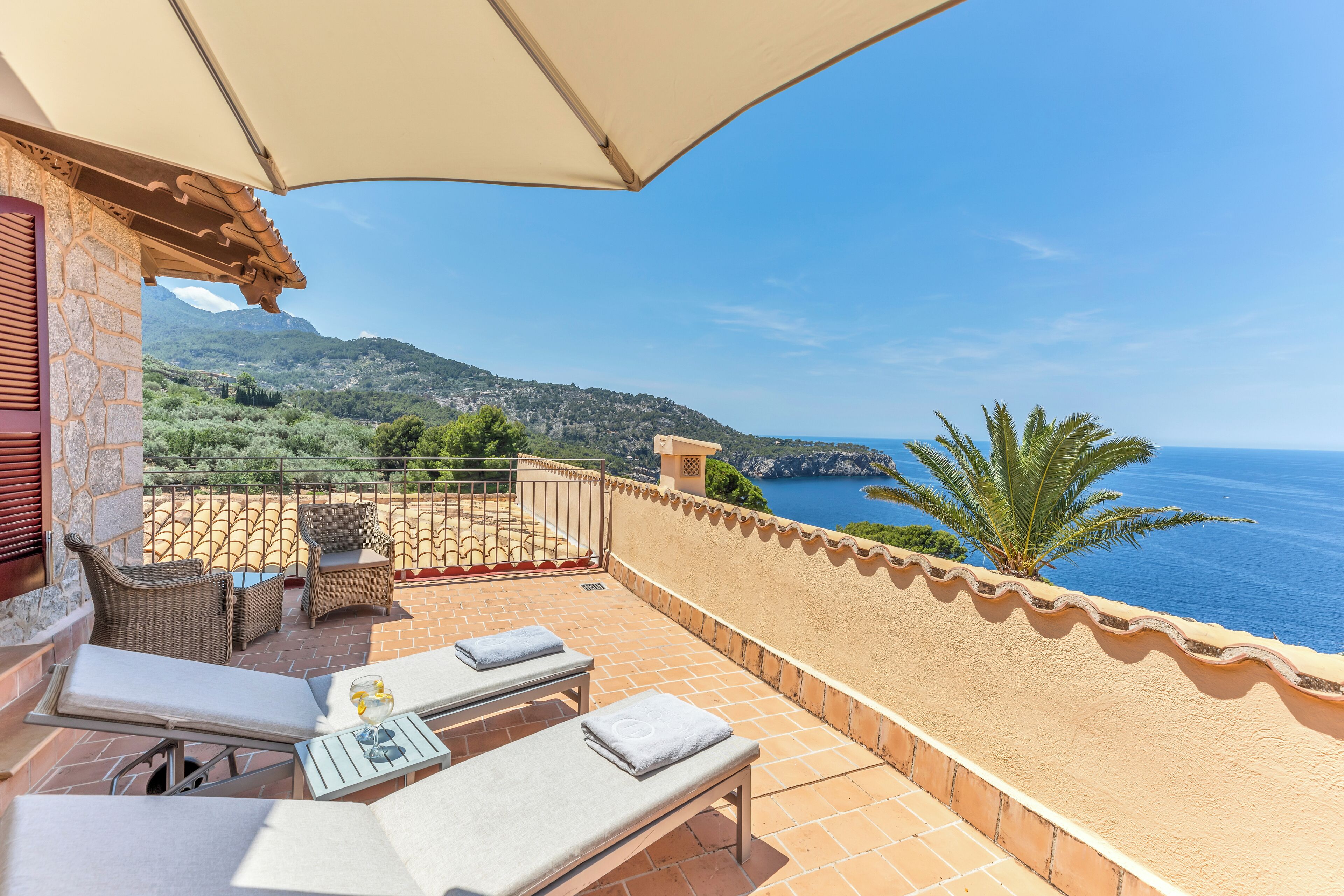 A terrace with two lounge chairs and a table, overlooking a coastal landscape with a clear view of the sea and distant mountains.