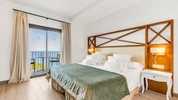 Quarto de hotel com cama, mesa de cabeceira, abajur, vista do mar através de uma varanda e cabeceira de madeira.