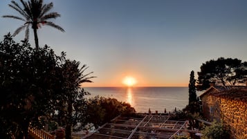 Vista de un atardecer costero con una terraza que da al mar, rodeada de vegetación y un edificio de piedra.