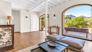 Una habitación amplia con suelo de madera, un sofá, una mesa y vistas a un jardín a través de grandes ventanales.