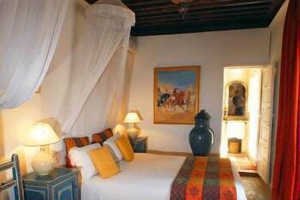 A bedroom with a bed, two bedside tables with lamps, a red chair, a vase, and a painting on the wall.