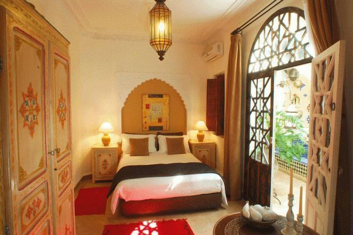 A traditional room with a bed, two bedside tables with lamps, a decorative arched wall, and a view through a lattice window.