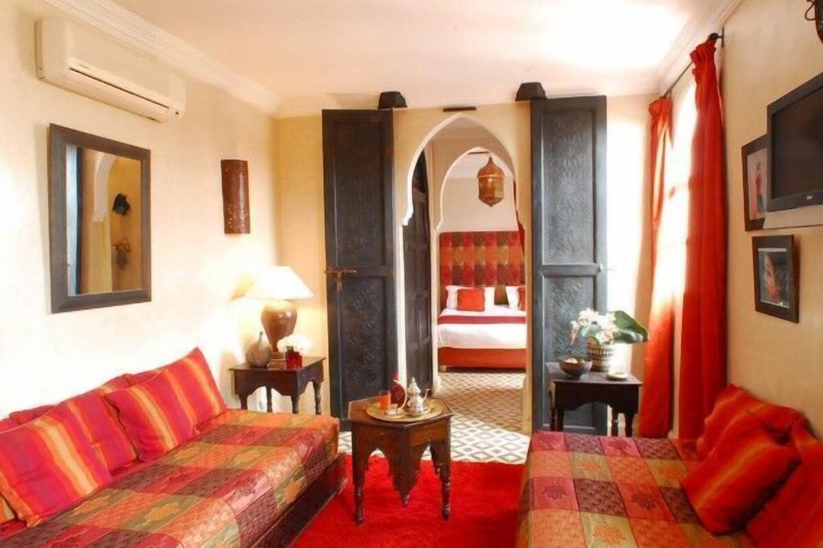 A hotel room with two sofas, a small table with a lamp, and a bed with red bedding.
