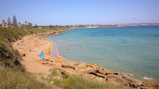 A beach with clear blue water, rocky shoreline, and people sunbathing. - Perla del Sole (Syracuse)